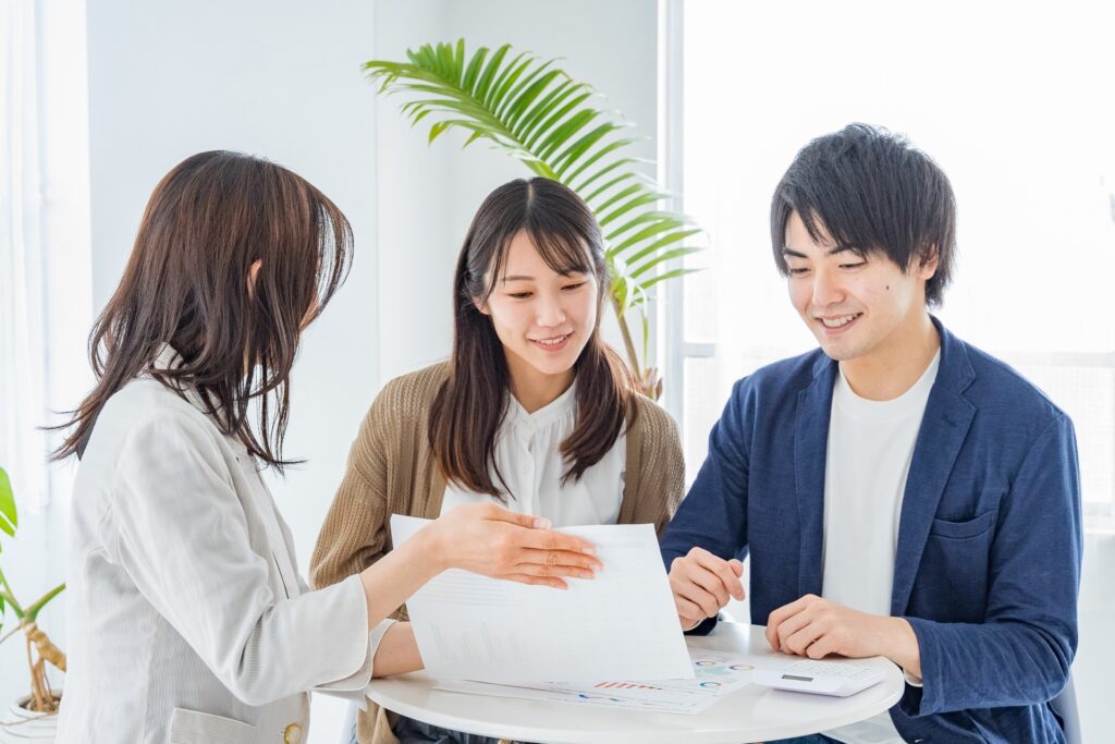 明るいオフィスのテーブルで、白いジャケットを着た女性カウンセラーがグラフ資料を指さしながら、笑顔の若い夫婦にライフプランや各種手続きの相談に乗っている様子。